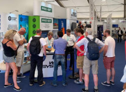 Audience members standing watching a demonstration at stand in boat show exhibition hall.