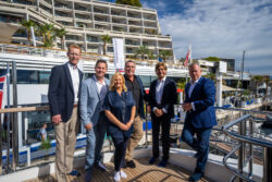 Six people standing on top open deck of motor yacht in a marina on a sunny day.