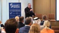 View of backs of heads in an audience that is listening to a speaker delivering a talk at a presentation.