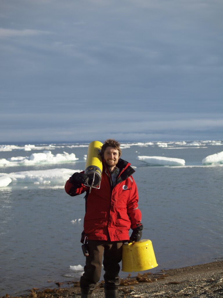 Bearded man on beach in red jacket carrying yellow research gear. The waves crash behind him.