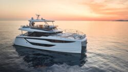 A luxury catamaran with four people on board, floating in the sea off the coast with orange/yellow sky of sunset in the background.